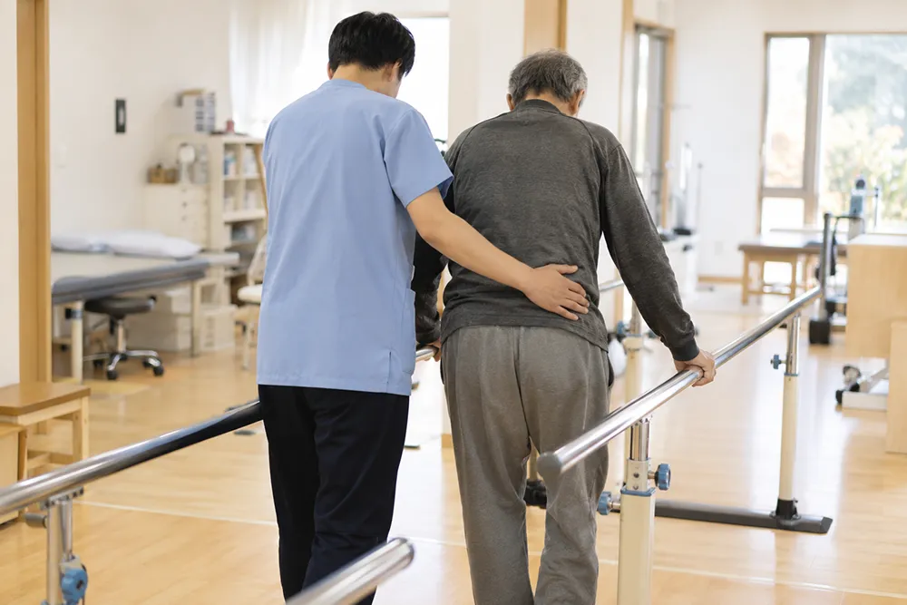 平行棒につかまって歩行練習をする方と、腰を支える理学療法士